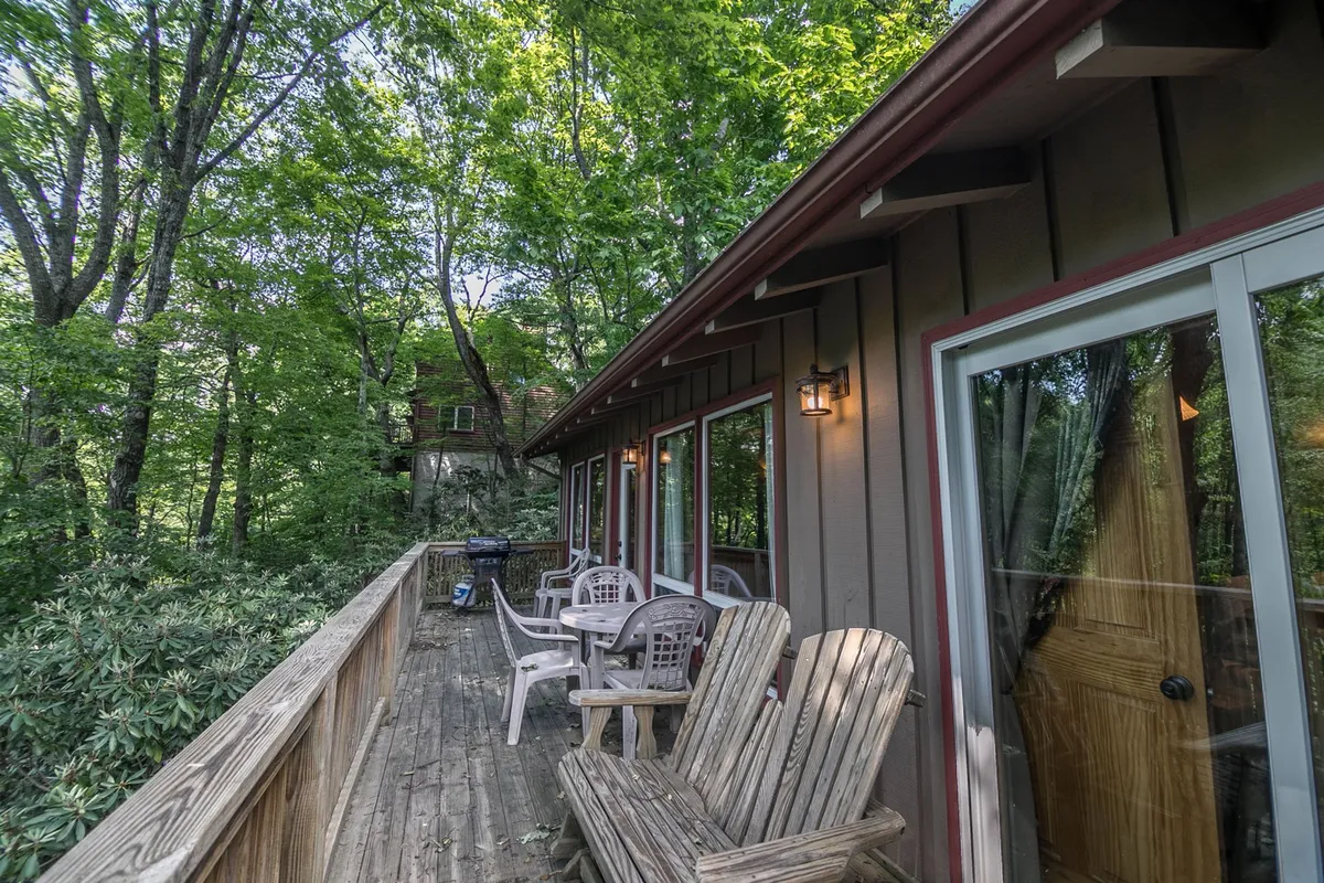 A comfortably furnished covered deck nestled among tall trees, featuring outdoor seating and a gas BBQ grill. The deck is accessed directly from the main living area, creating a seamless flow between indoors and out, and is set up for relaxation, grilling, and savoring mountain air with pets or loved ones.