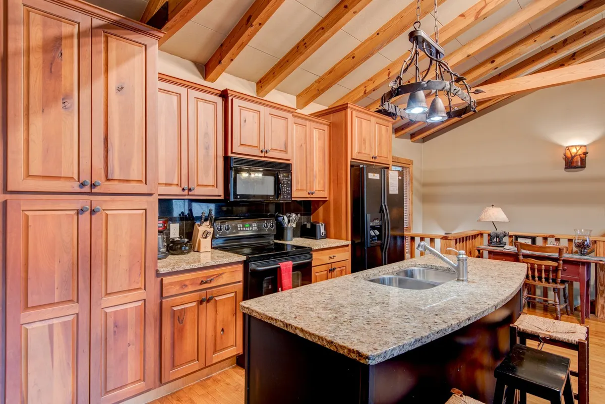 A warm, welcoming kitchen with a granite island, wooden cabinetry, exposed wooden beams, and modern appliances. The space is set up for both meal preparation and entertaining, with inviting lighting and a homey, rustic feel.