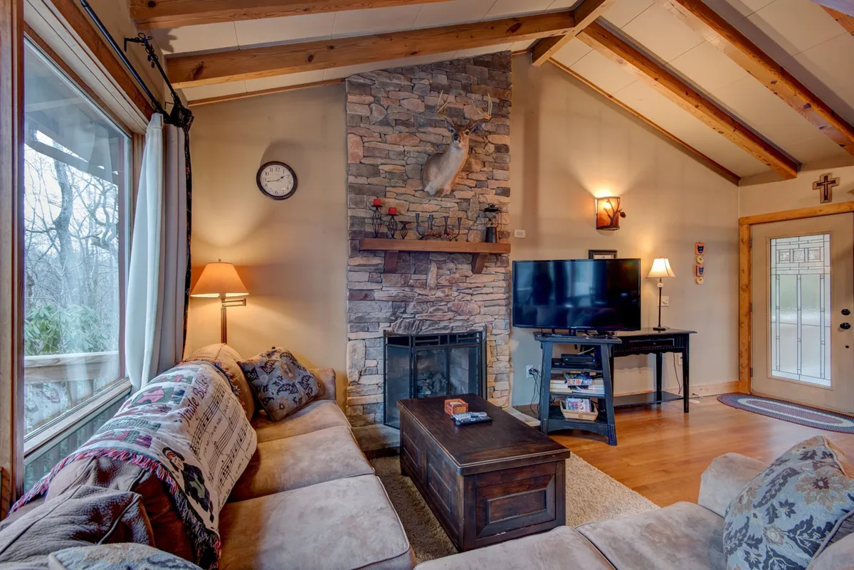 The hero image showcases a cozy living room with a striking stacked stone gas log fireplace as the centerpiece, surrounded by plush sofas and accented with rustic exposed wooden beams. Natural light filters in, creating a warm and inviting atmosphere perfect for unwinding. The setting evokes a sense of relaxation, comfort, and mountain charm.