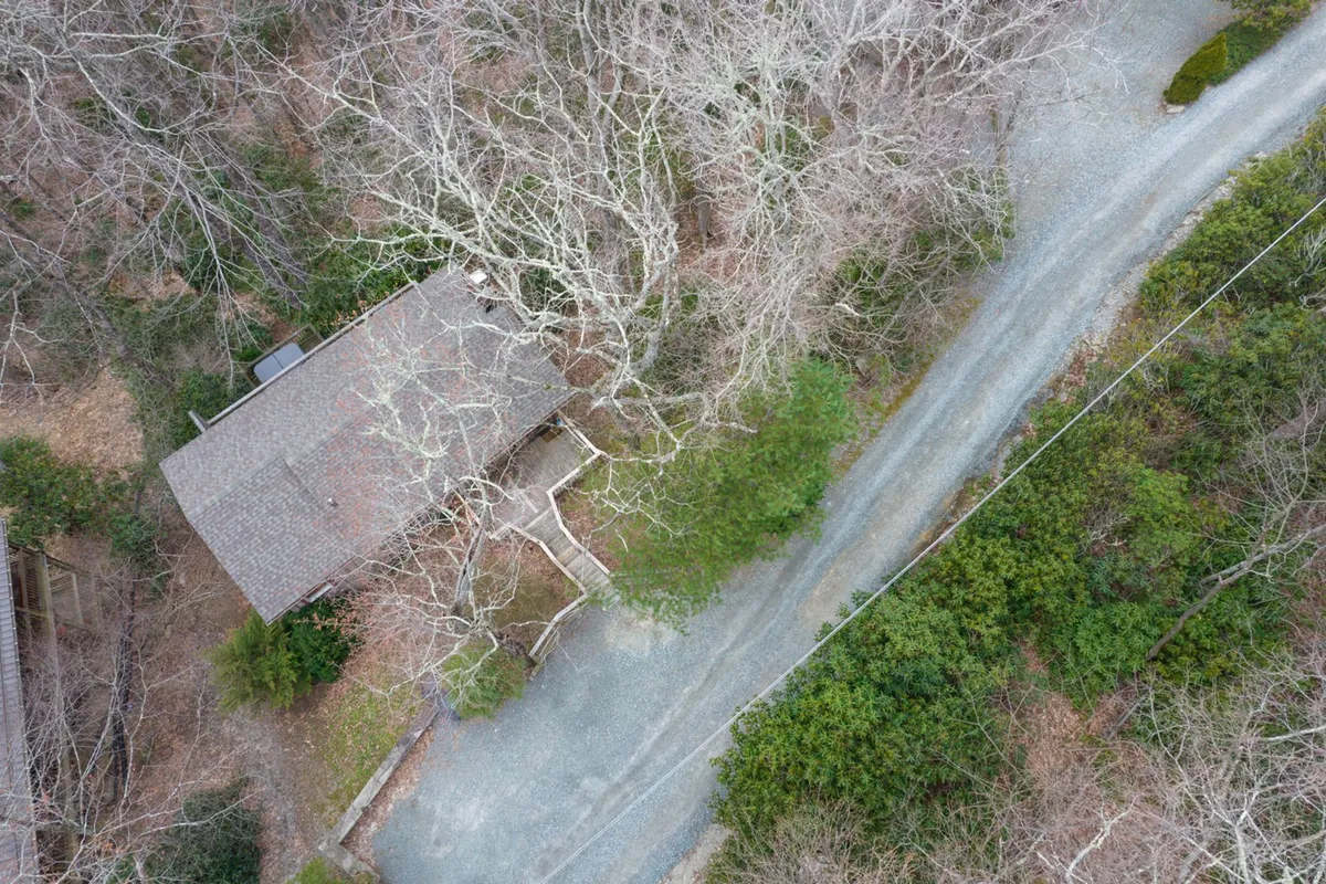 The home is pictured with its classic wooden exterior, a winding gravel road, and dense trees surrounding the property. This wide-angle perspective shows the property within its natural mountain setting, illustrating easy access to adventure and the peacefulness of the retreat.