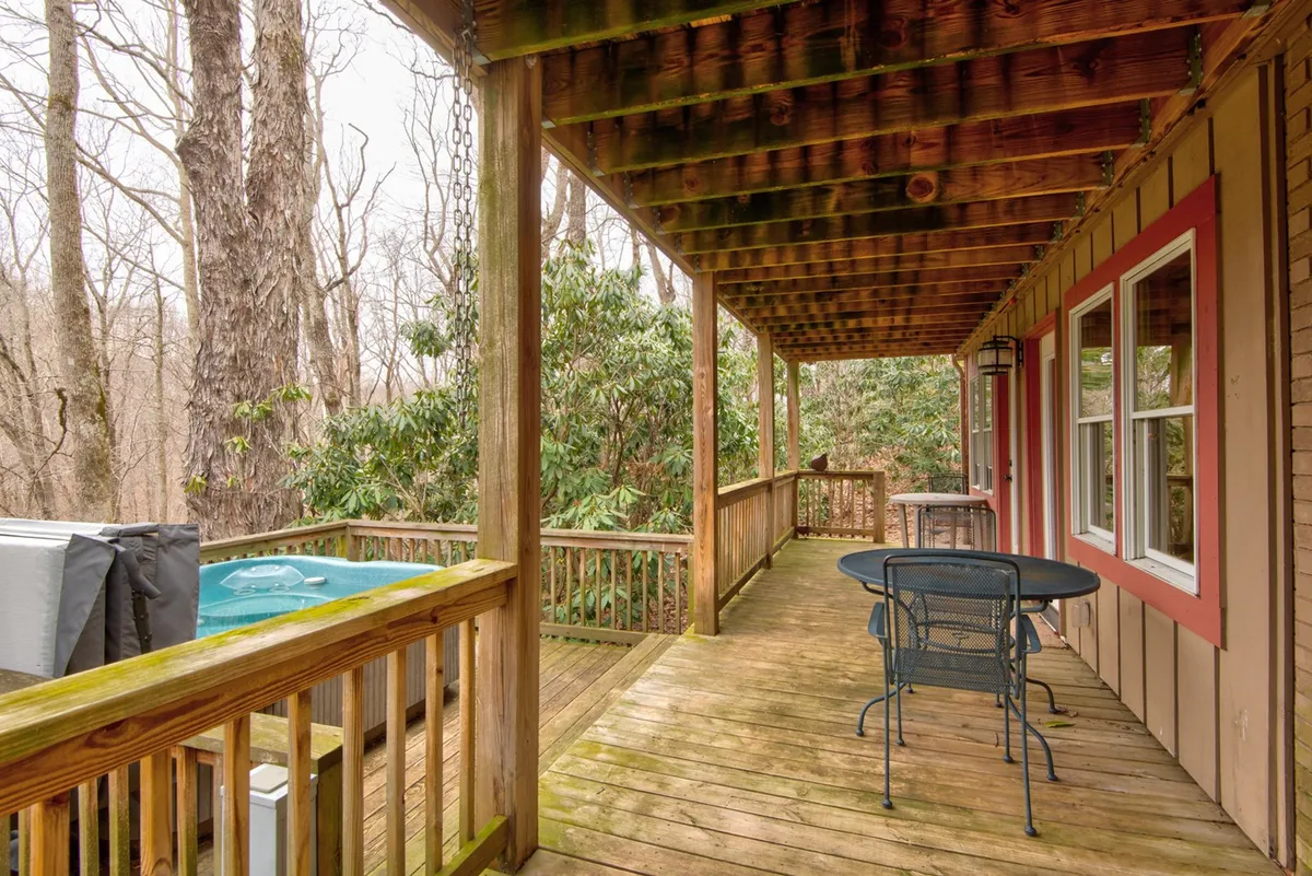 This exterior image shows a peaceful wooden deck nestled among trees, featuring a private hot tub, a table and chairs, and views of the surrounding forest. The scene captures a sense of seclusion and tranquility, perfect for morning coffee or an evening soak under the stars.
