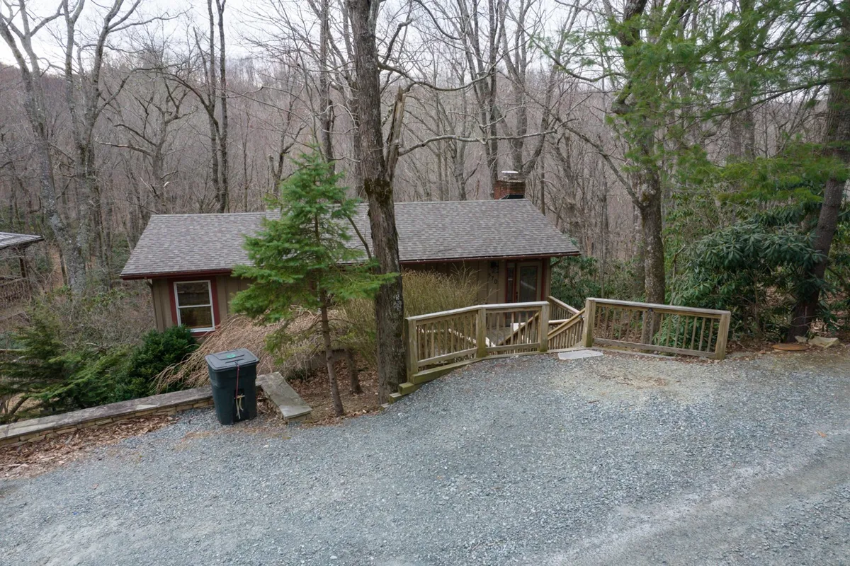 Set against a lush mountain backdrop, this image showcases the exterior of a wooden home surrounded by trees, a gravel driveway, and a pathway leading to the entrance. The natural setting enhances the feeling of a peaceful, secluded retreat.