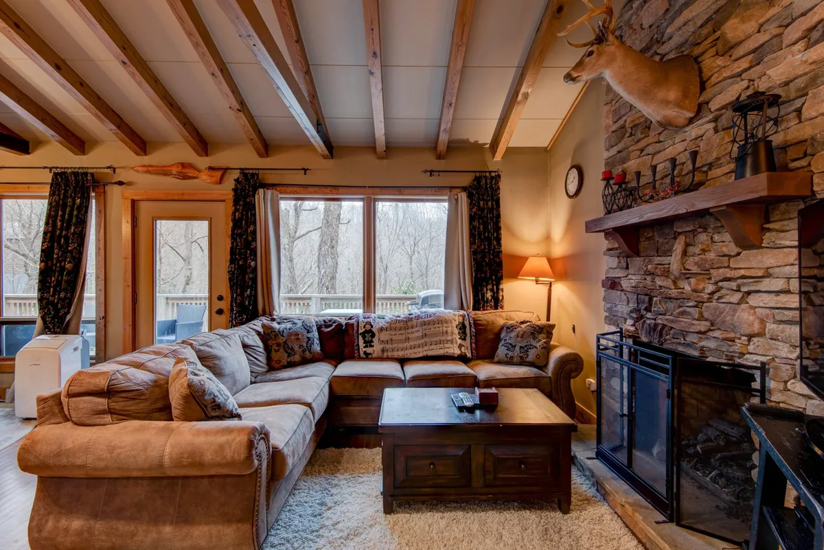 A spacious and inviting main living area featuring a large sectional sofa, a striking stacked-stone fireplace, expansive windows that flood the space with natural light, exposed wooden beams overhead, and mountain-inspired décor including a decorative deer head above the mantle. The setting radiates warmth and encourages gathering, with ample seating for family and friends.