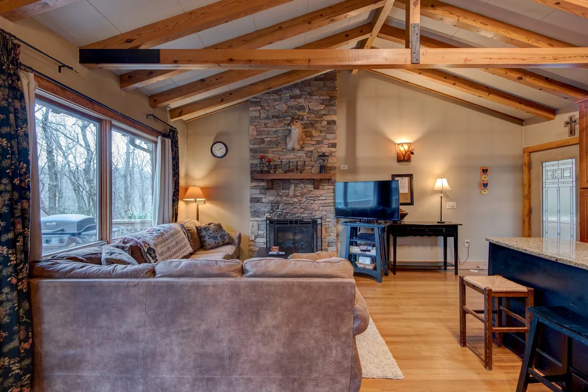 A spacious, inviting interior with a plush sofa facing a stone fireplace and a large HDTV, with expansive windows revealing forest views. The scene radiates warmth, comfort, and the perfect setting for movie nights or peaceful relaxation by the fire.