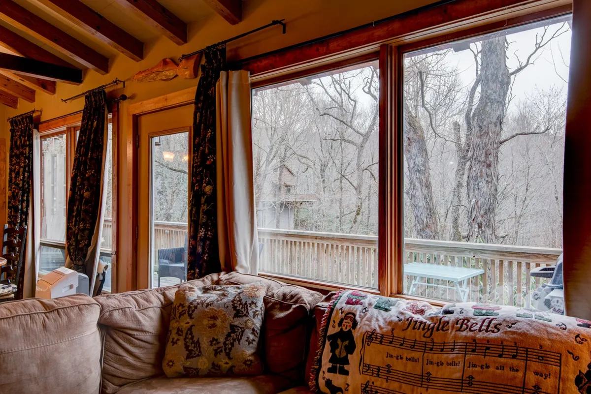 This interior image features a cozy sofa set against large windows that offer a view of the surrounding trees. Decorative pillows and the abundance of natural light create a warm, inviting space ideal for both relaxation and observing nature from indoors. The layout suggests plenty of room for people and pets to lounge comfortably.