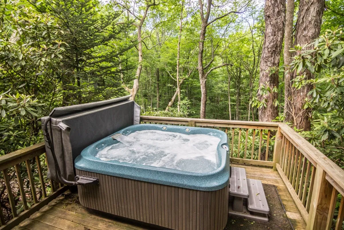A serene exterior shot of the lower wooden deck featuring a bubbling hot tub surrounded by lush greenery and tranquil forest views. The hot tub is the focal point, emphasizing relaxation in a natural setting.