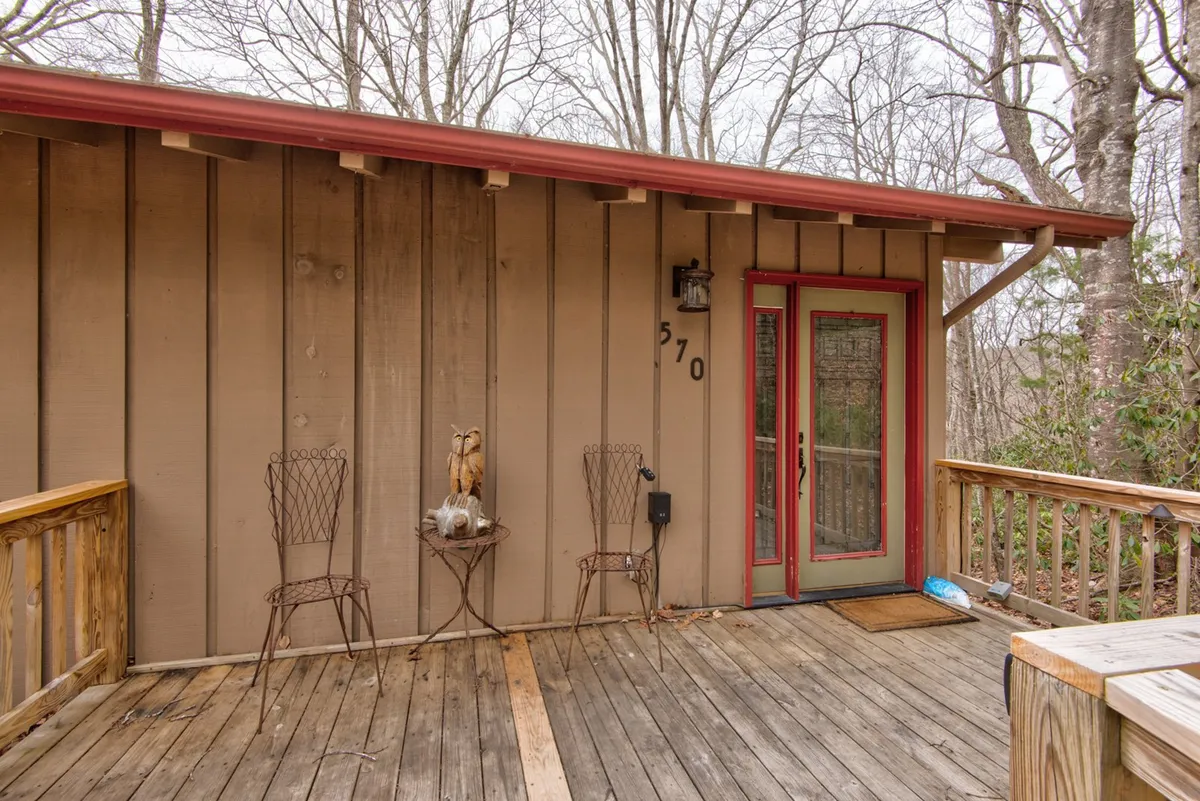 An exterior shot of the wooden deck, complete with rustic chairs and a decorative table, positioned near the house's red door. The setting is cozy and welcoming, offering a picturesque space for enjoying nature, sipping coffee, or unwinding with pets beside you.